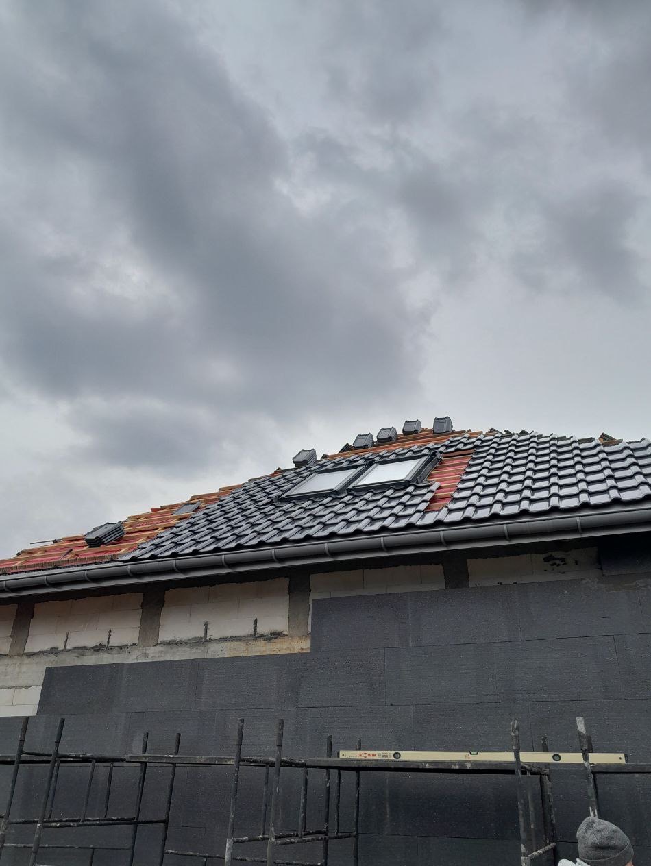 Montaż dachówek ceramicznych na dachu z oknami połaciowymi, widoczne rusztowania i ocieplenie budynku styropianem grafitowym. Pochmurne niebo.