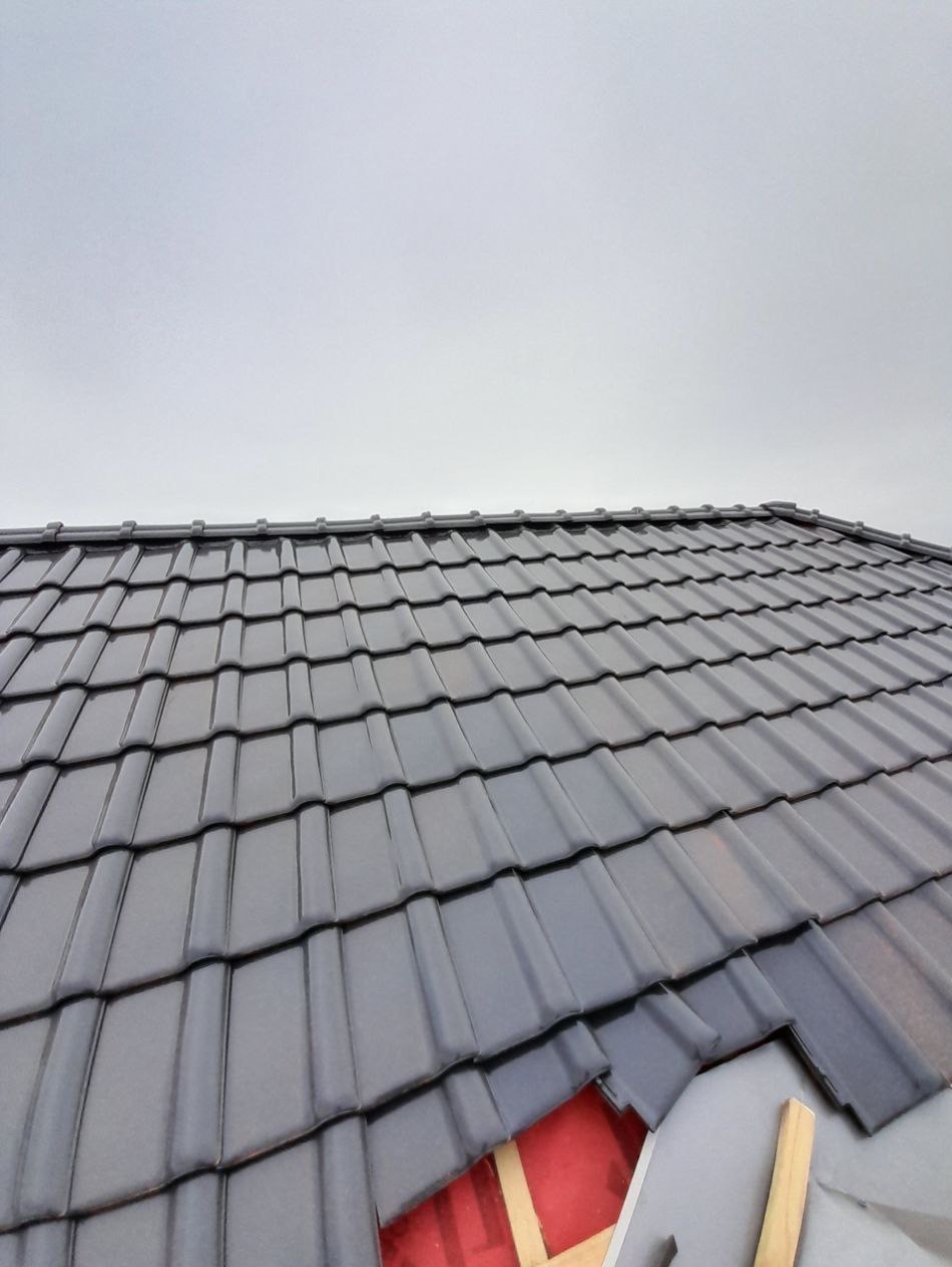 Fragment dachu krytego błyszczącą, grafitową dachówką ceramiczną. Widoczne detale konstrukcji dachu i czerwona membrana w miejscu braku dachówek. Pochmurne niebo w tle.