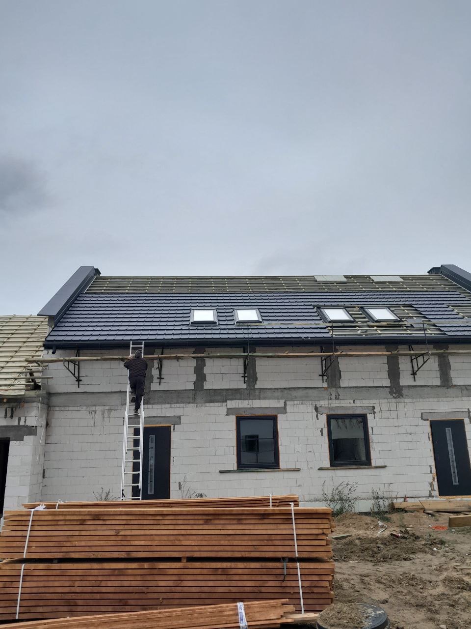 Montaż ciemnej dachówki na nowym domu z oknami dachowymi. Na drabinie dekarz, w tle szare niebo. Na dole drewniane belki.