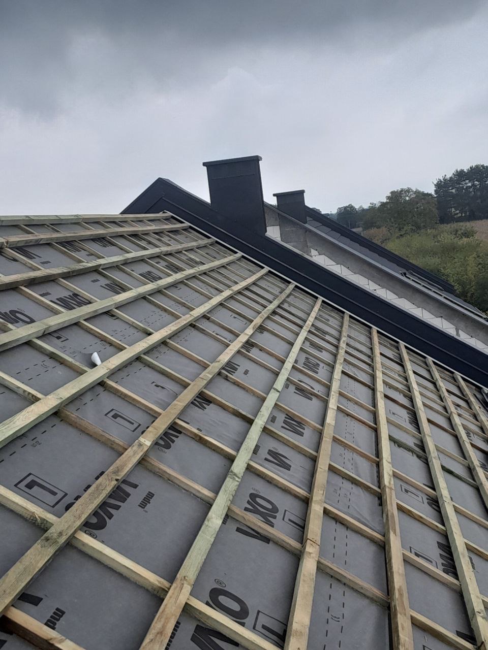 Konstrukcja dachu z łatami i membraną, czarne kominy w tle, pochmurne niebo. Widok z dołu ukazuje detale przygotowania pod pokrycie.