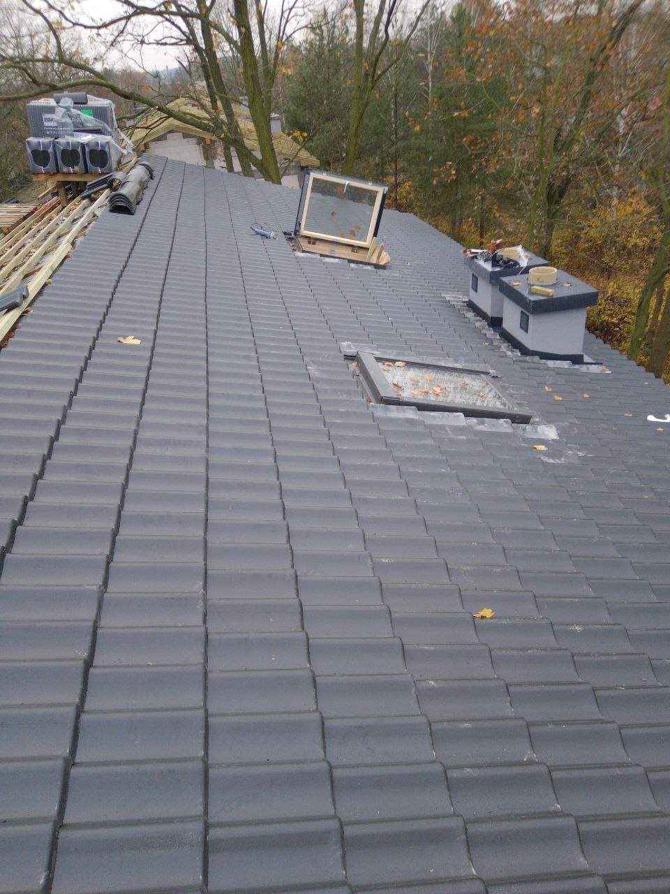 Szary dach z dachówki ceramicznej z oknami dachowymi, częściowo zamontowanymi. Na dachu widoczne narzędzia i zapas dachówek. Jesienne drzewa w tle.