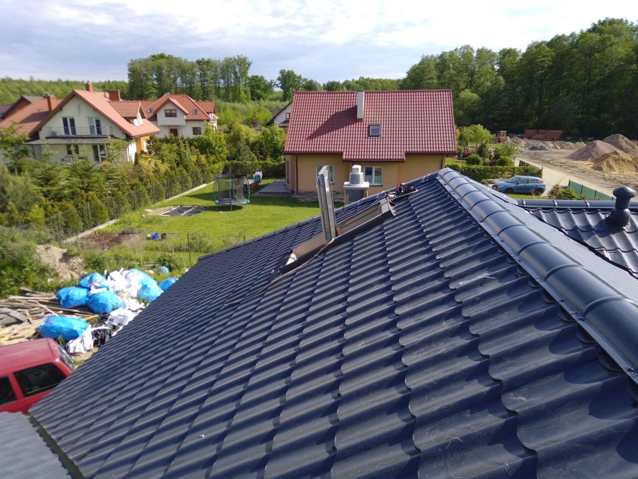 Ukończony dach z ciemnej dachówki ceramicznej, widok z góry na tle zabudowy jednorodzinnej i zieleni. Widoczny komin i fragmenty obróbek blacharskich.