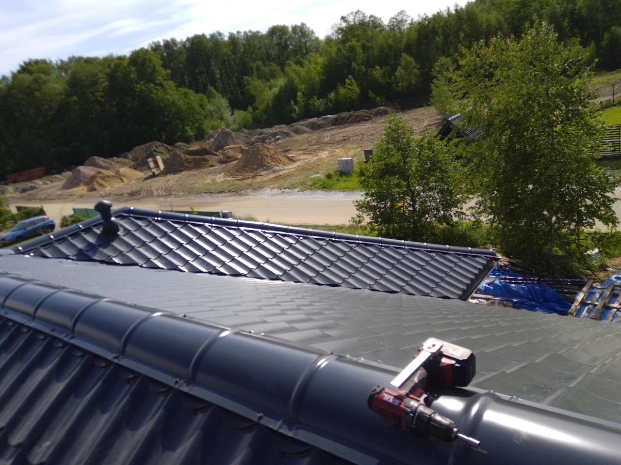 Ciemnoszary dach z dachówki, z widoczną wkrętarką na kalenicy. W tle drzewa i plac budowy.