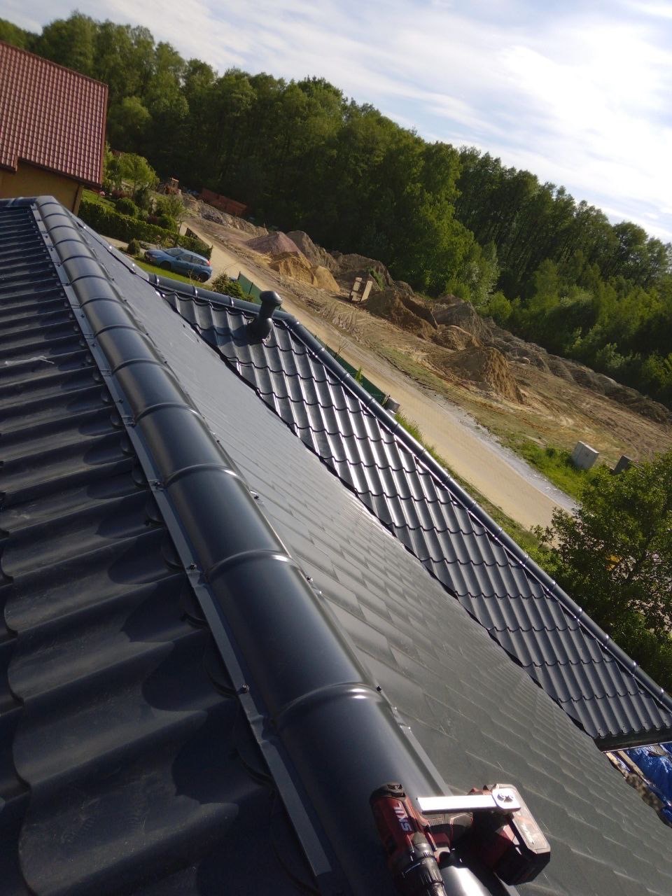 Ciemnoszara dachówka ceramiczna z zamontowaną rynną w kolorze antracytowym. Widoczne narzędzie do montażu. W tle drzewa i błękitne niebo.