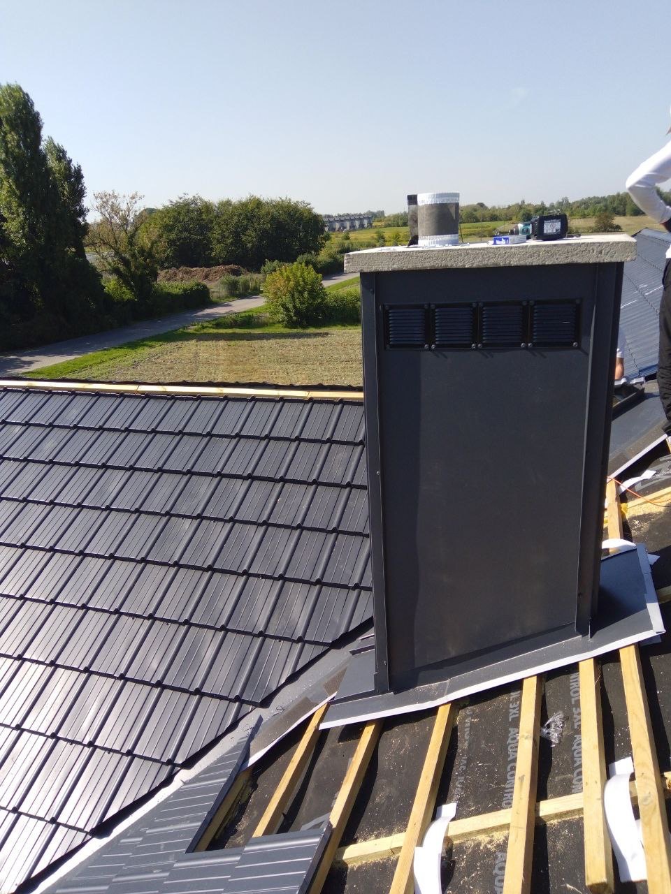 Ciemnoszara dachówka ceramiczna na połaci dachu w trakcie montażu, widoczny komin wentylacyjny i elementy konstrukcji drewnianej. Słoneczny dzień.