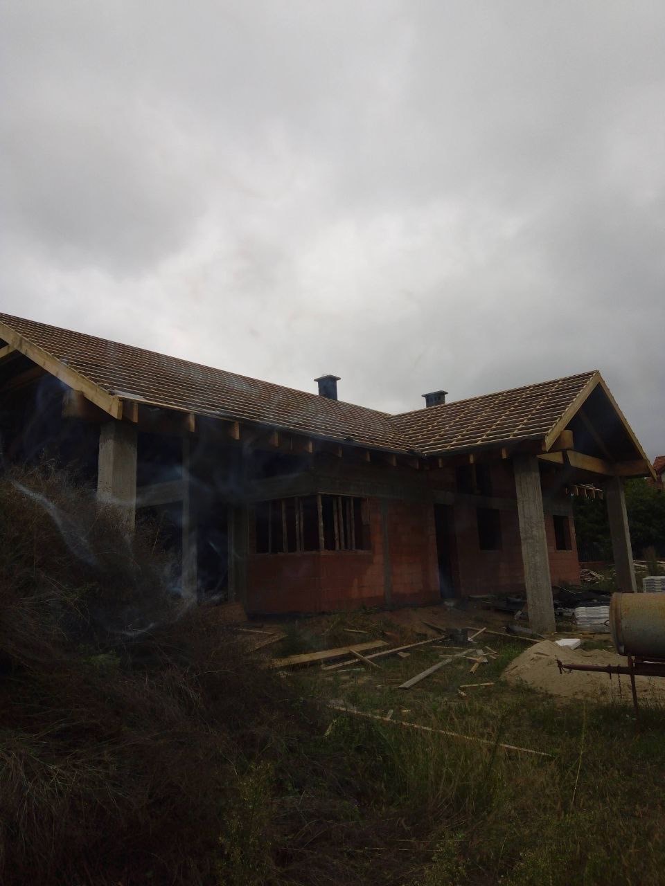 Budowa domu jednorodzinnego w trakcie krycia dachówką ceramiczną. Widoczne elementy konstrukcji dachu i kominy, na pierwszym planie pozostałości po budowie.