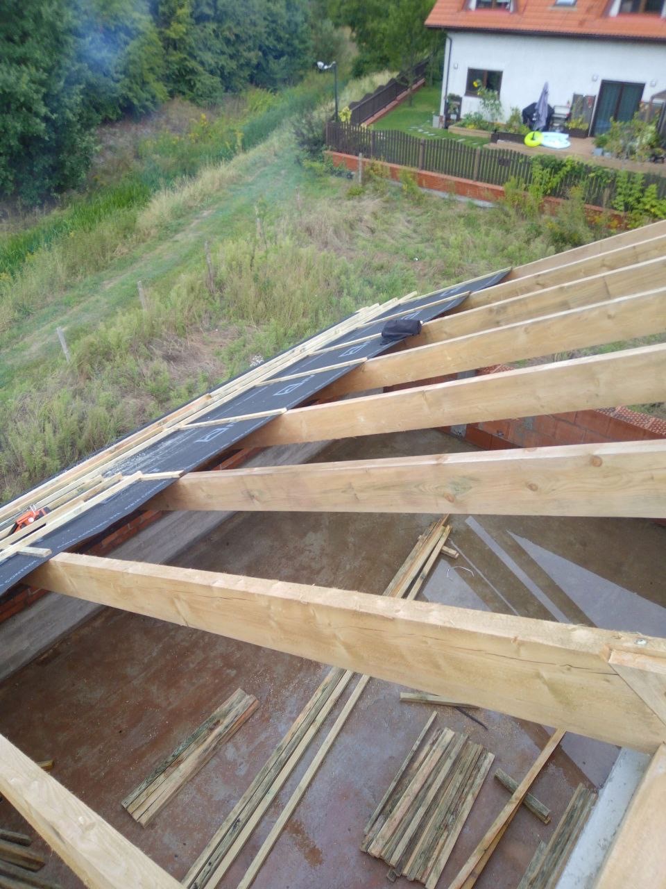 Konstrukcja drewniana dachu w trakcie budowy, widok z góry na krokwie i membranę, w tle dom jednorodzinny i zieleń.