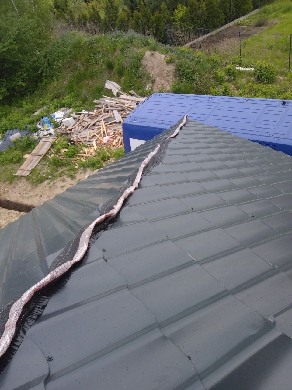 Szary dach z zamontowaną membraną kalenicową, widok z góry na tle zieleni i niebieskiego kontenera. Detal dachu po montażu, zabezpieczenie przed wilgocią.