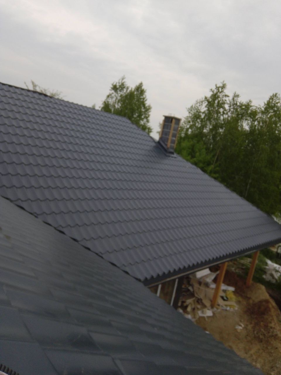 Szary dach z metalowej dachówki z kominem w tle drzew i pochmurnego nieba, widziany z góry pod kątem. Widoczny fragment drugiego dachu.