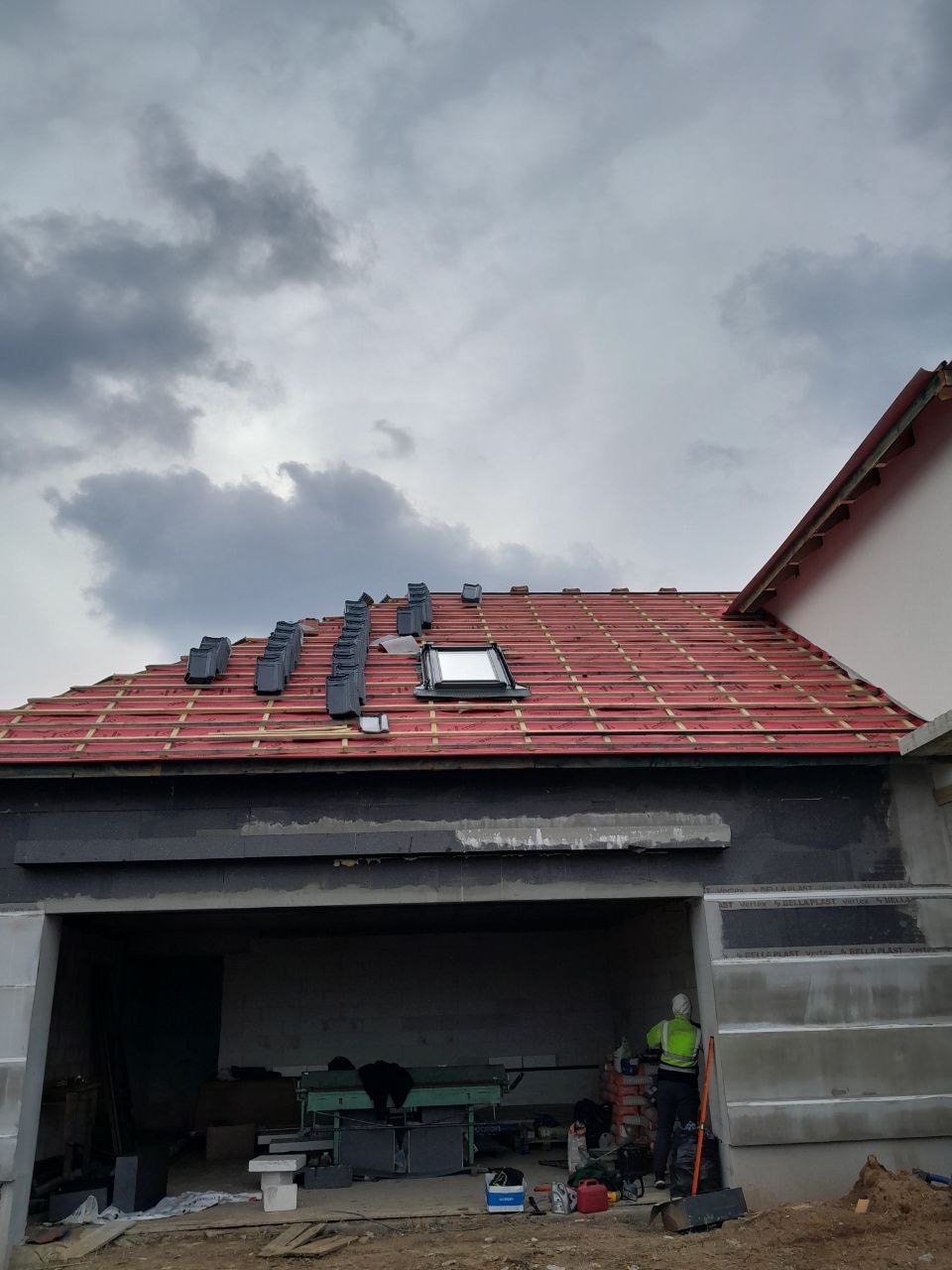 Częściowo pokryty dach z oknem dachowym i ułożonymi dachówkami w rzędach. Widoczny fragment budynku w trakcie budowy i pracownik w odblaskowej kamizelce.