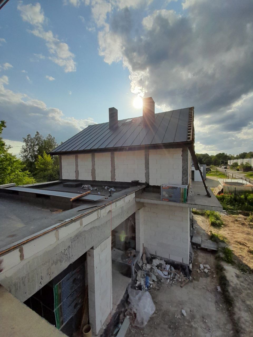 Surowy stan domu z szarą, metalową konstrukcją dachu i dwoma kominami, widok z góry, słoneczny dzień.