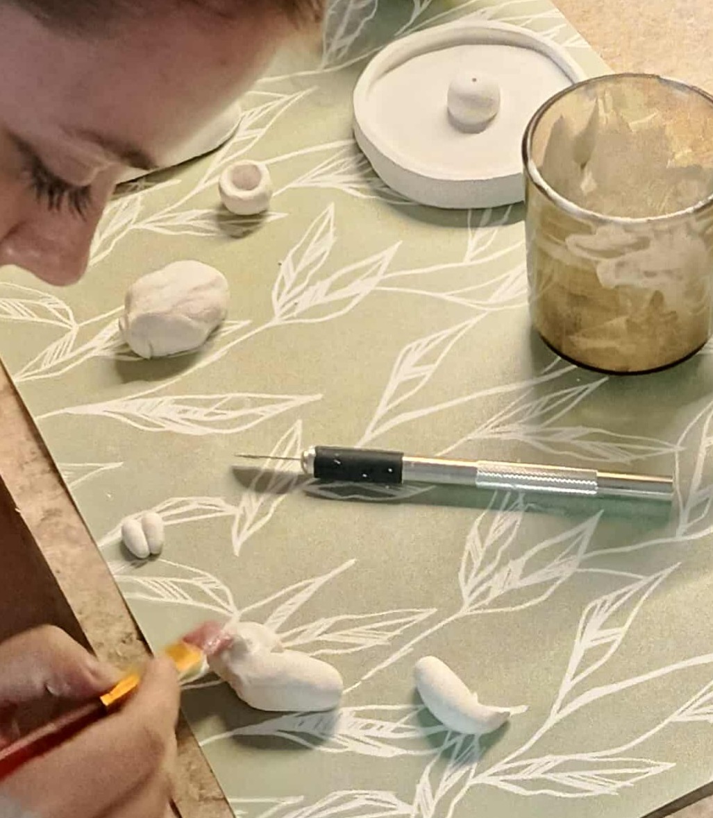 Osoba maluje pędzelkiem białą figurkę z gliny na tle zielonego podkładu z liśćmi. Obok leżą narzędzia i inne elementy gliniane.