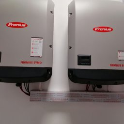 Mateusz Żakowski - Dwa inwertery Fronius Symo zamontowane na ścianie, podłączone przewodami, z widocznymi naklejkami informacyjnymi i logo producenta. Instalacja fotowoltaiczna.