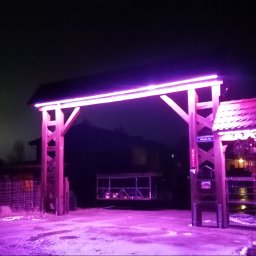 Mateusz Żakowski - Podświetlona na fioletowo drewniana brama wjazdowa z oświetleniem LED, widoczna nocą. Na słupku tabliczka z nazwą ulicy. W tle zarys domu i drzew.