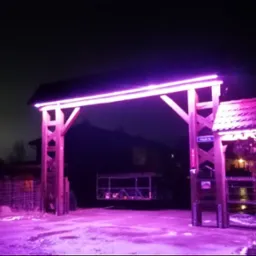 Podświetlona na fioletowo drewniana brama wjazdowa z oświetleniem LED, widoczna nocą. Na słupku tabliczka z nazwą ulicy. W tle zarys domu i drzew.