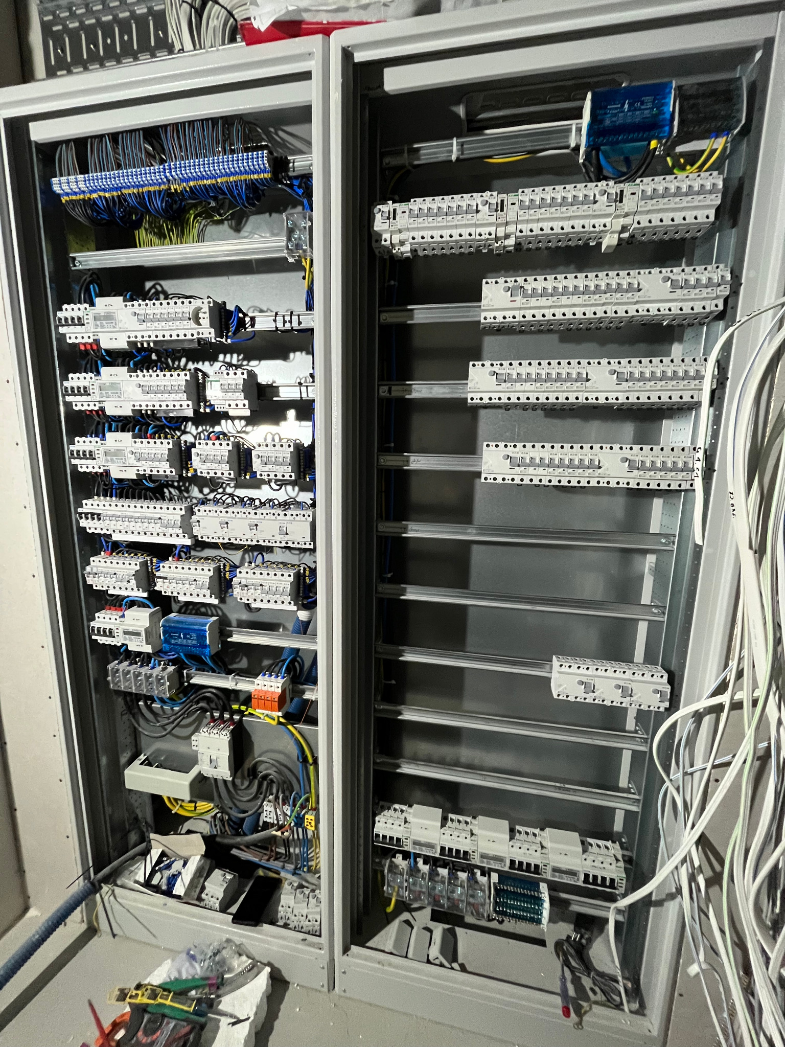 Rozdzielnia elektryczna w metalowej obudowie, z licznymi przewodami i zabezpieczeniami, widok z bliska, z narzędziami na podłodze.