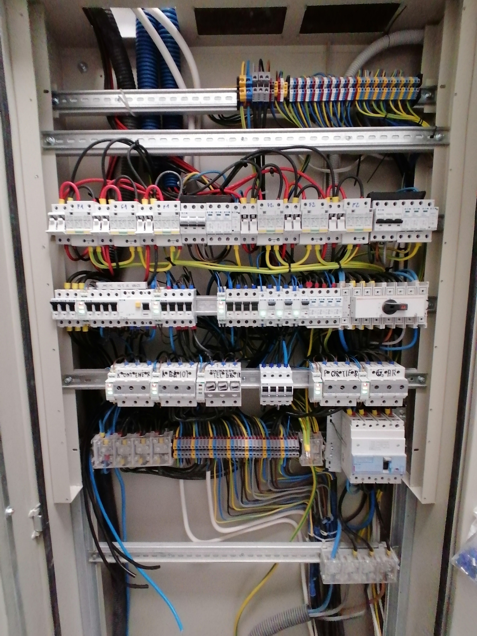 Rozdzielnica elektryczna z licznymi zabezpieczeniami, przewodami o różnych kolorach i listwami montażowymi wewnątrz obudowy. Widoczne detale instalacji.