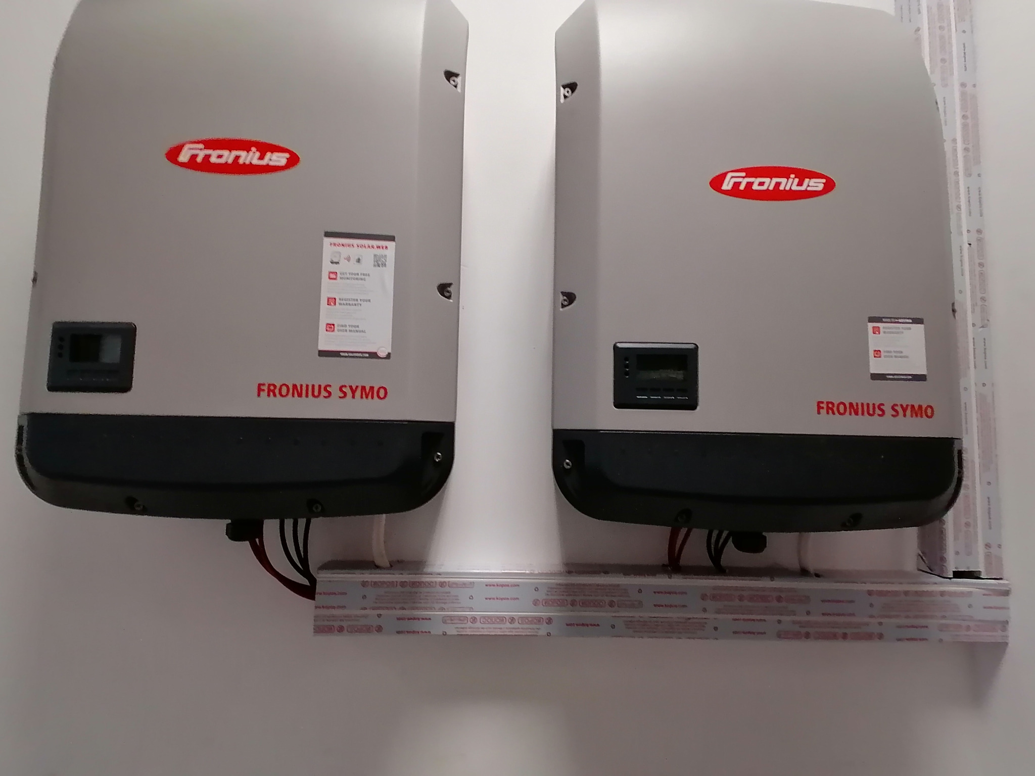 Dwa inwertery Fronius Symo zamontowane na ścianie, podłączone przewodami, z widocznymi naklejkami informacyjnymi i logo producenta. Instalacja fotowoltaiczna.