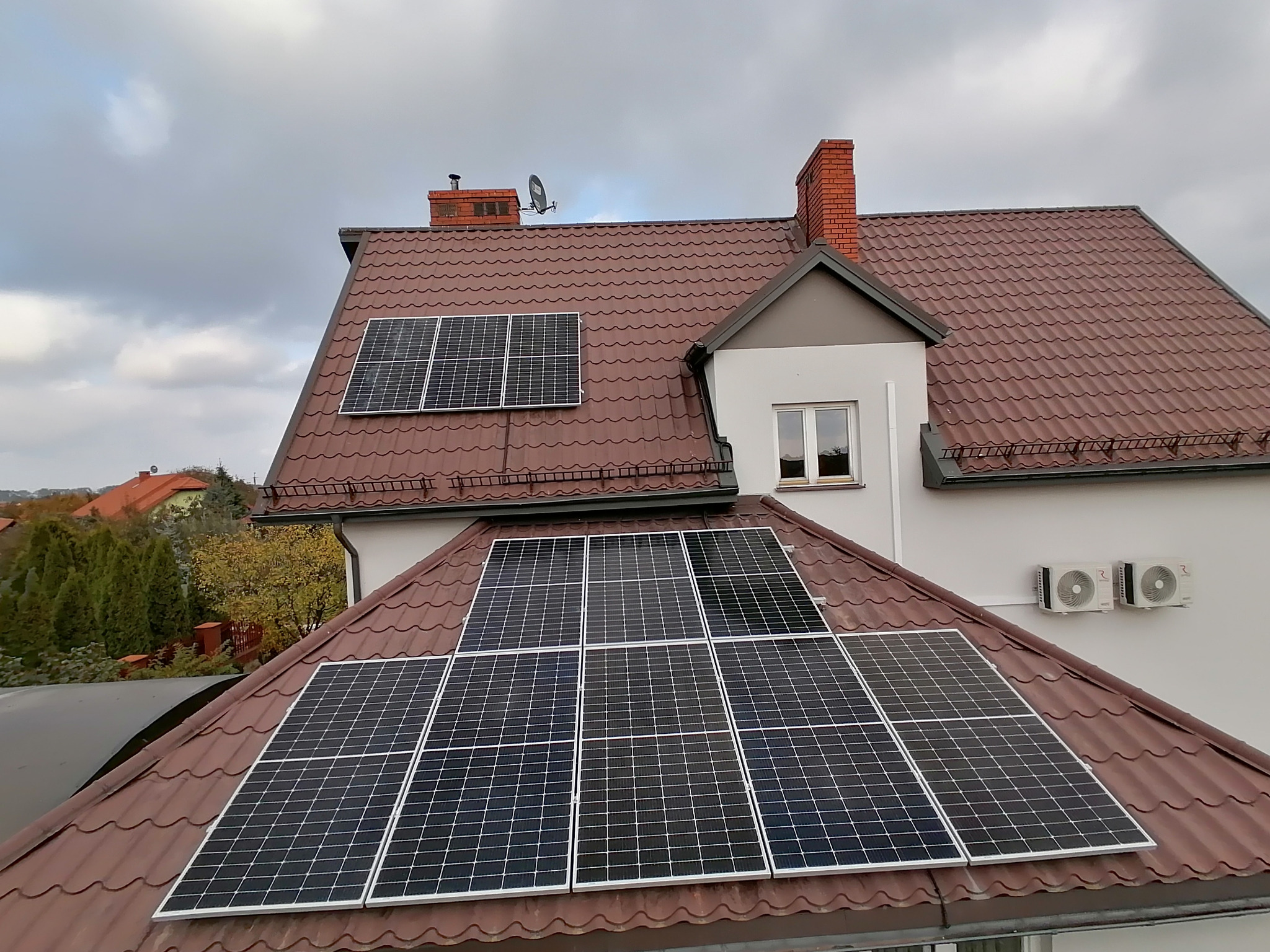 Panele fotowoltaiczne na brązowym dachu domu jednorodzinnego, widoczne również klimatyzatory na ścianie i komin z cegły. Pochmurne niebo w tle.