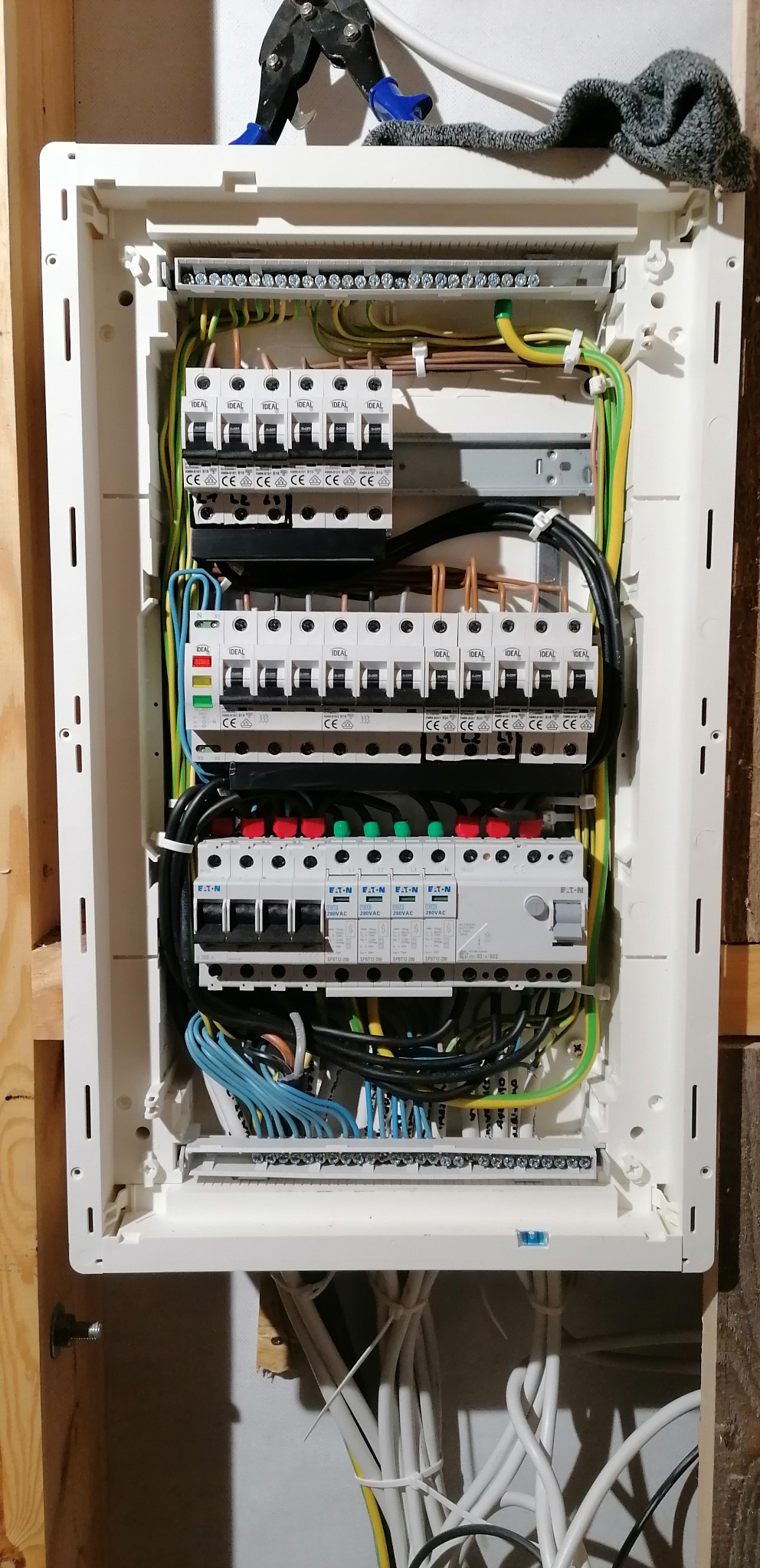 Rozdzielnica elektryczna w obudowie z widocznymi bezpiecznikami, przewodami i listwami zaciskowymi. Nad nią leżą kombinerki i szmatka.