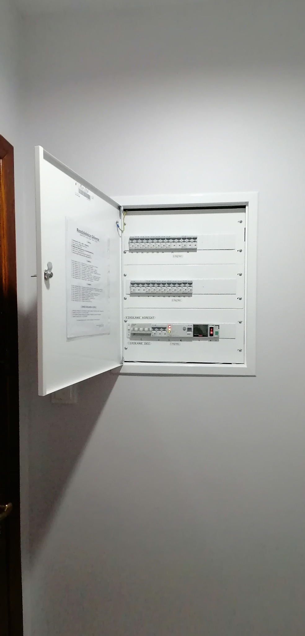 Otwarta, biała skrzynka rozdzielcza na ścianie, widoczne zabezpieczenia i schemat. Instalacja elektryczna wewnątrz budynku.