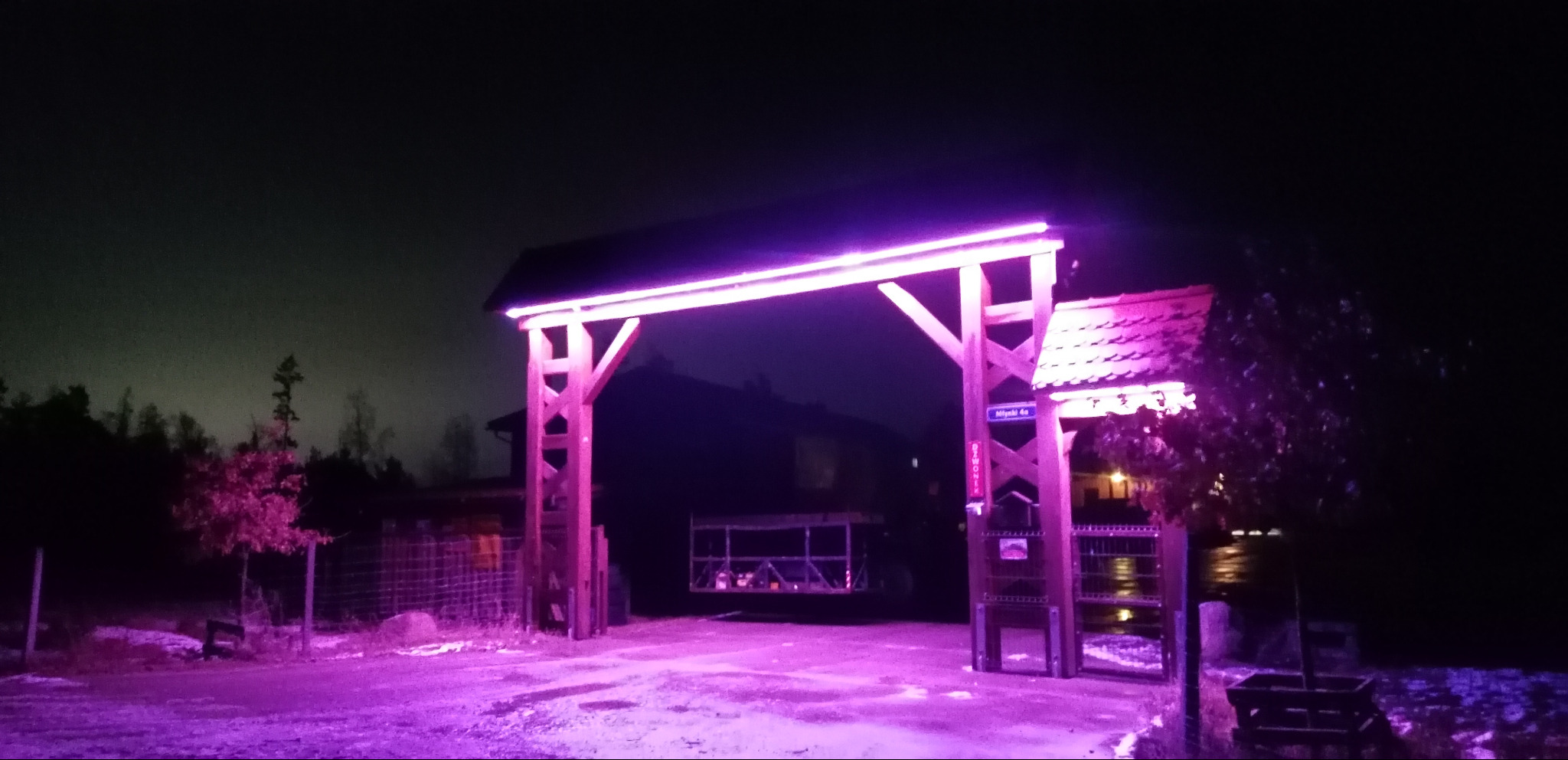 Podświetlona na fioletowo drewniana brama wjazdowa z oświetleniem LED, widoczna nocą. Na słupku tabliczka z nazwą ulicy. W tle zarys domu i drzew.