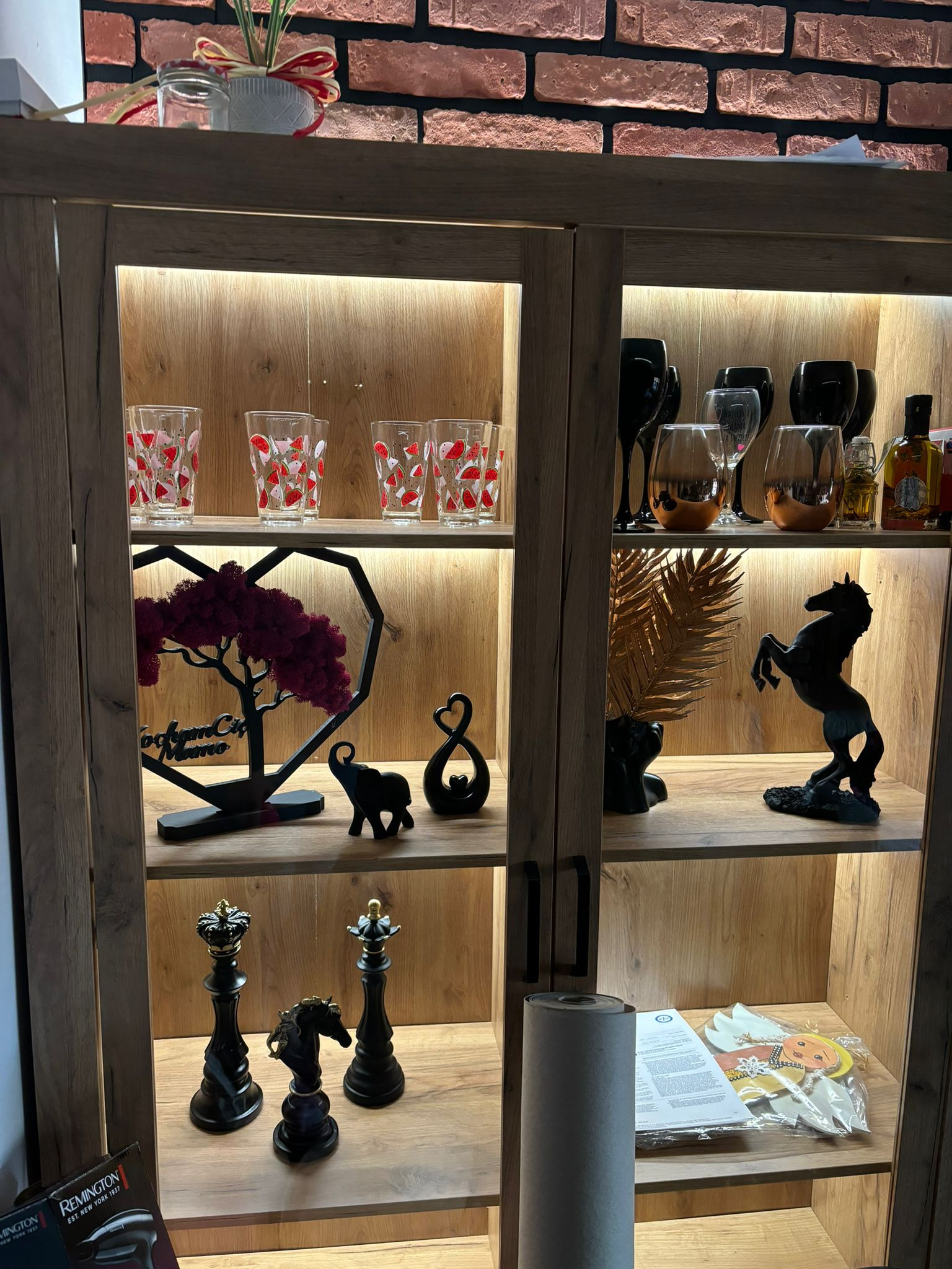 Oświetlona witryna z drewna z dekoracjami: szklanki w arbuzy, rzeźby koni, drzewko w sercu, figury słoni i szachów. Ceglana ściana w tle.