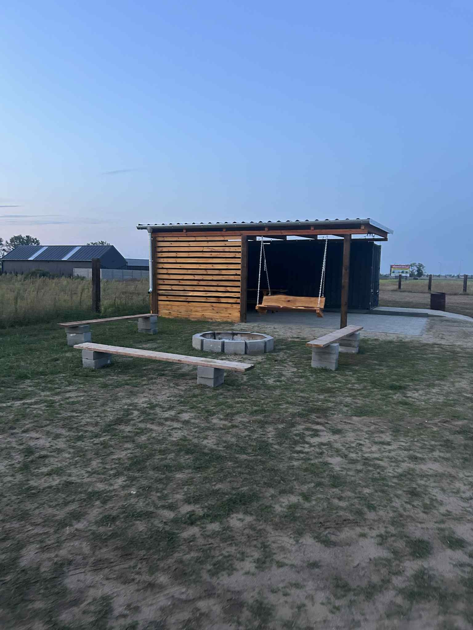 Miejsce na ognisko z drewnianą huśtawką i ławkami z betonowych bloków na trawiastym terenie, widoczne budynki w tle, oświetlone wieczornym światłem.