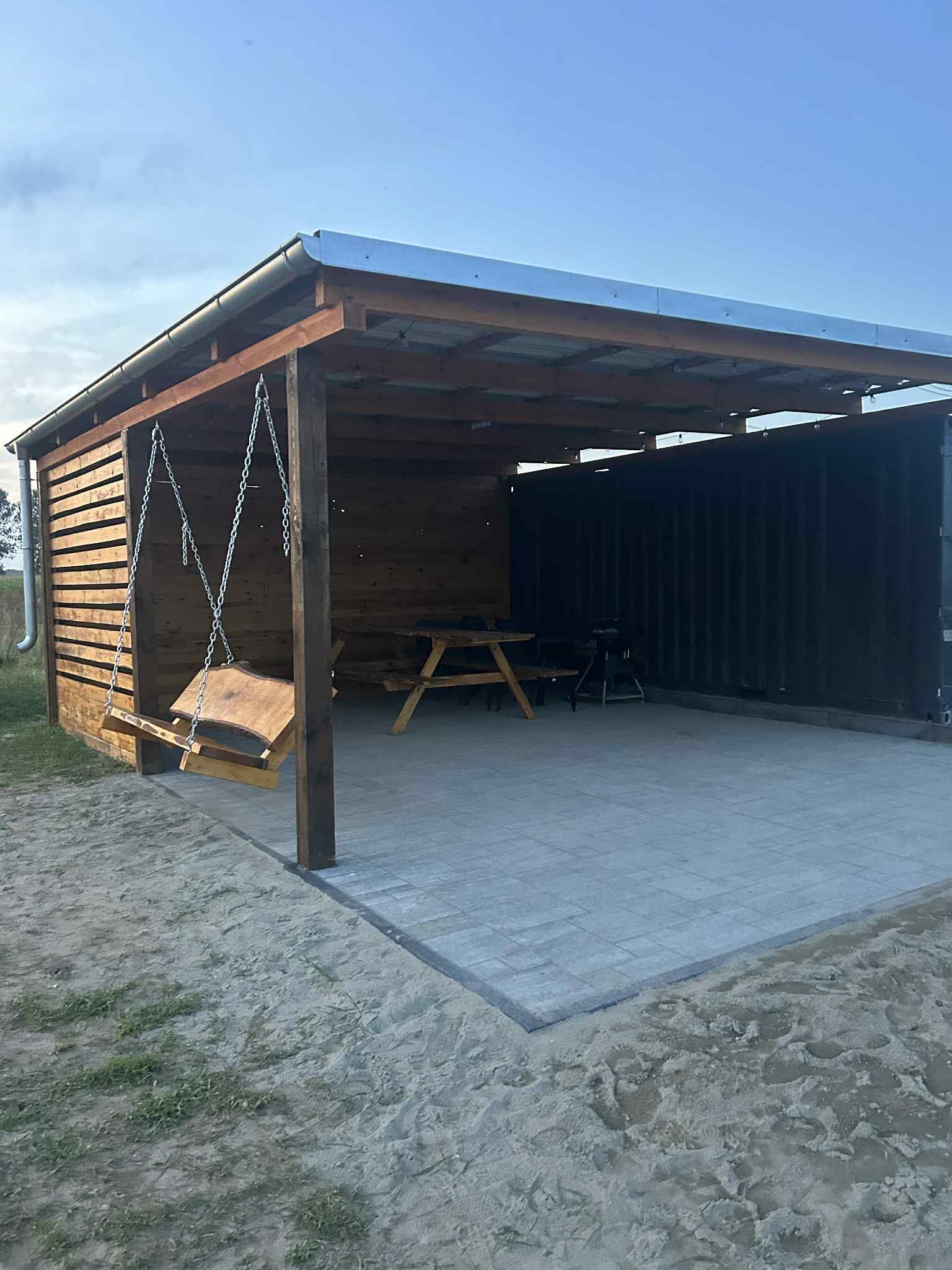 Zadaszona altana z drewnianą huśtawką i stołem piknikowym, wyłożona szarą kostką brukową. W tle widoczny kontener. Całość otoczona piaszczystym terenem.