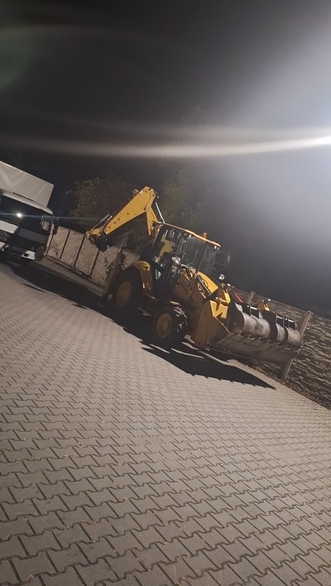 Żółta koparko-ładowarka na kostce brukowej nocą, oświetlona reflektorami. Z tyłu widoczna przyczepa transportowa. Perspektywa ukośna, podkreślająca gabaryty maszyny.