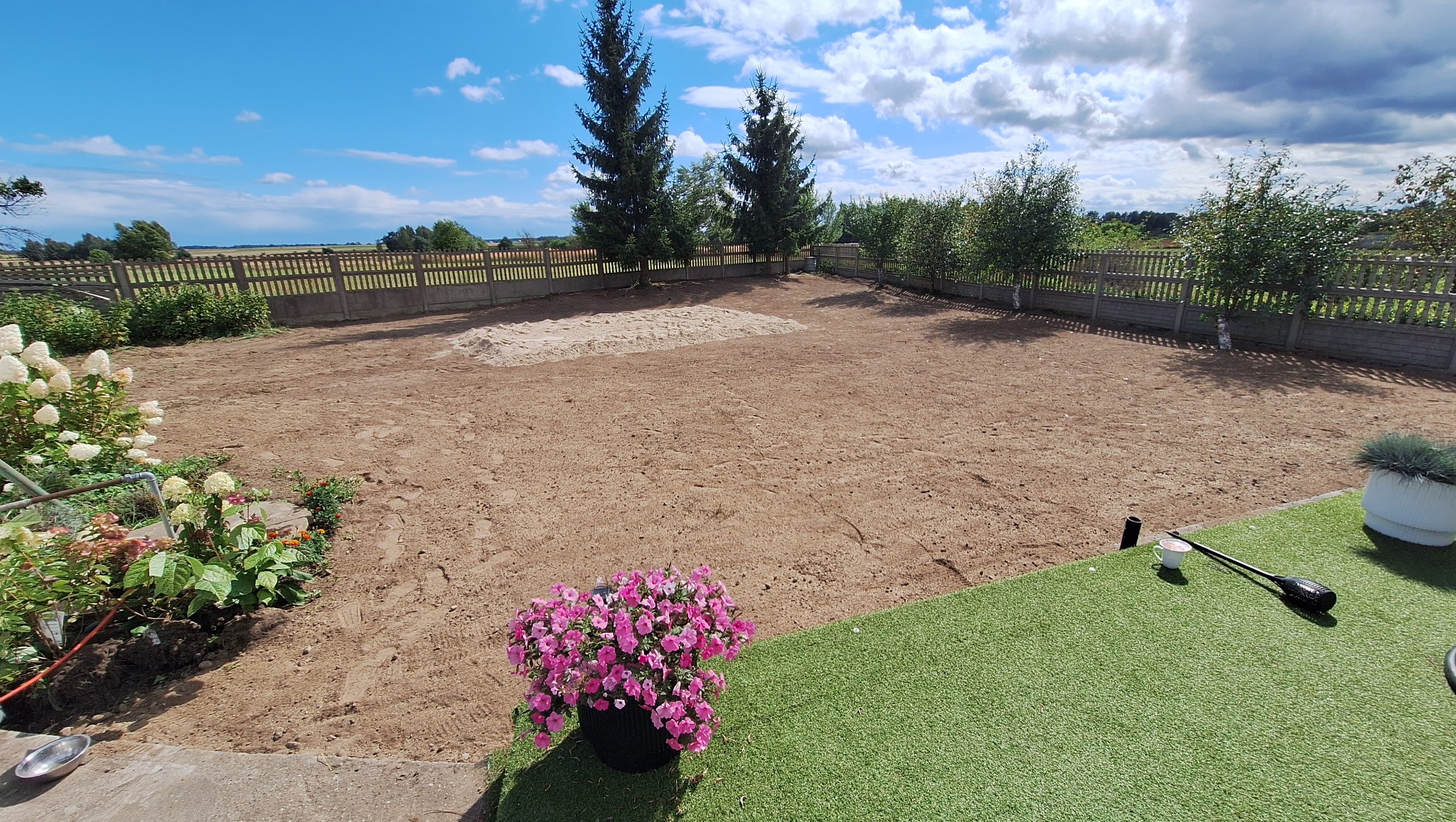 Przygotowany teren pod trawnik w Święciechowie. Wyrównana ziemia, widoczne ślady po pracy sprzętu, ogrodzenie w tle, kwiaty w donicach na pierwszym planie.