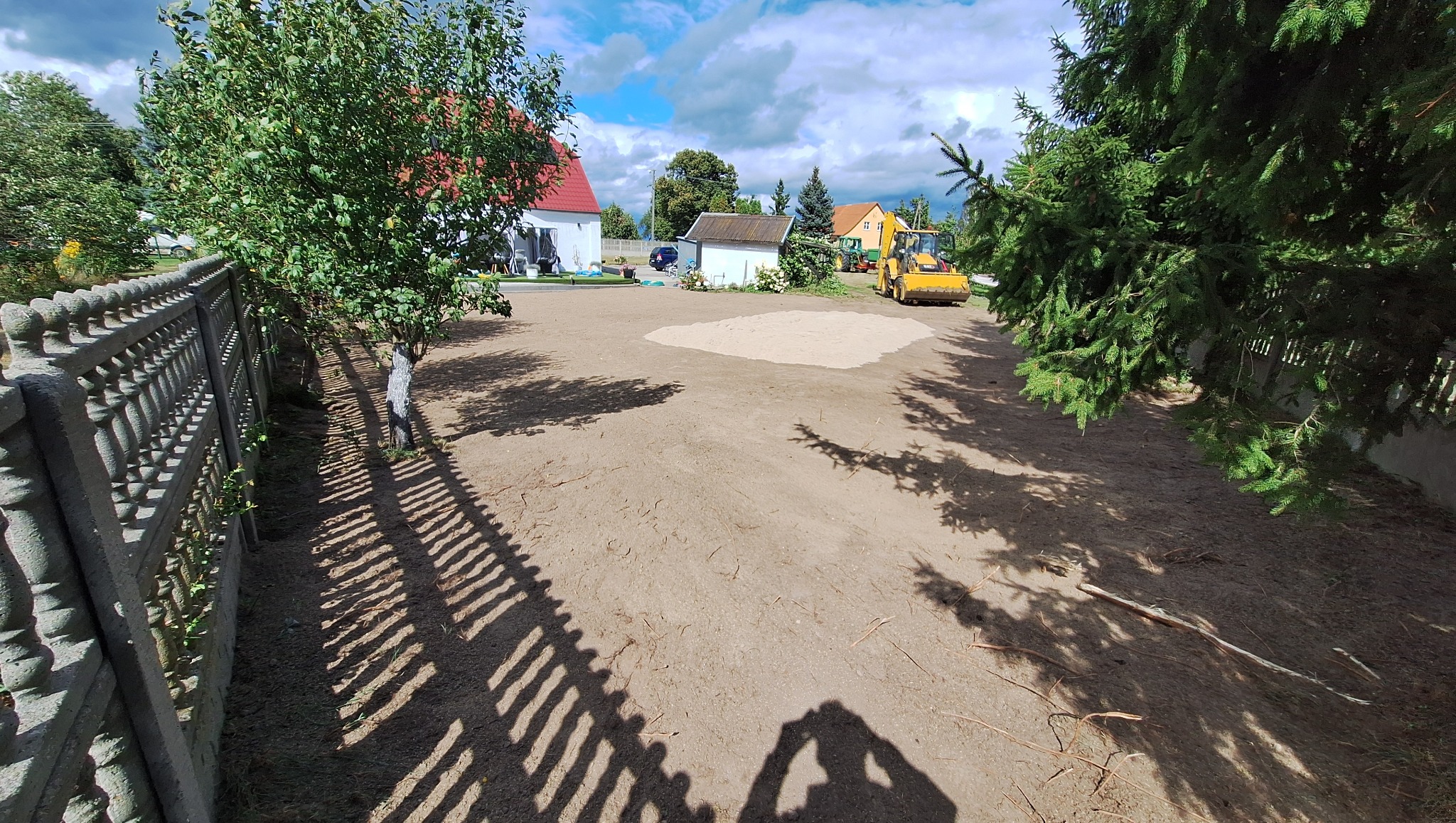 Żółta koparko-ładowarka na wyrównanym, piaszczystym terenie działki z widoczną szarą, betonową palisadą i drzewami w tle, w słoneczny dzień.