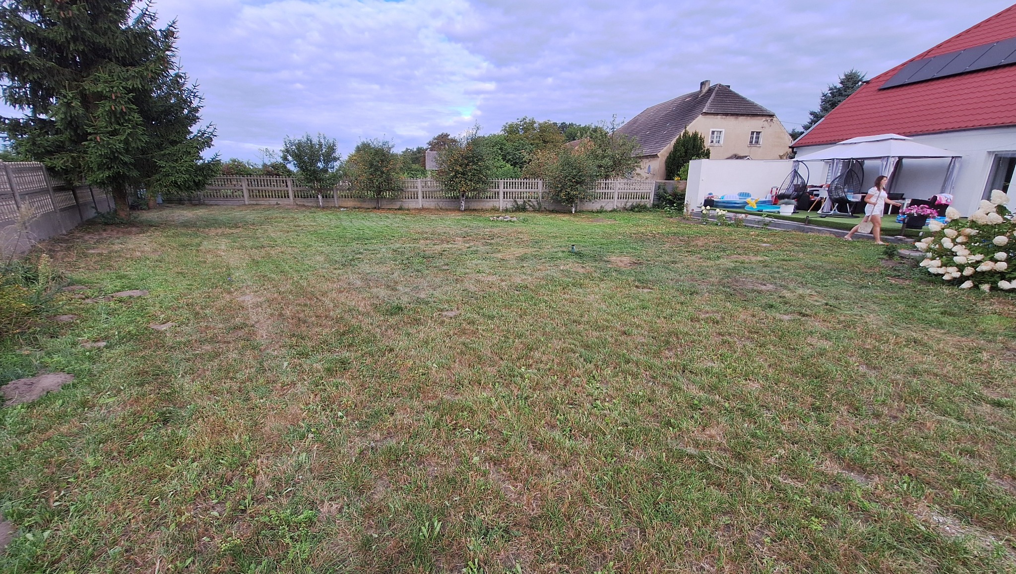 Rozległy, zaniedbany trawnik przed domem z widocznym ogrodzeniem i fragmentem budynku. Teren wymaga interwencji, widoczna wysoka trawa i nierówności.