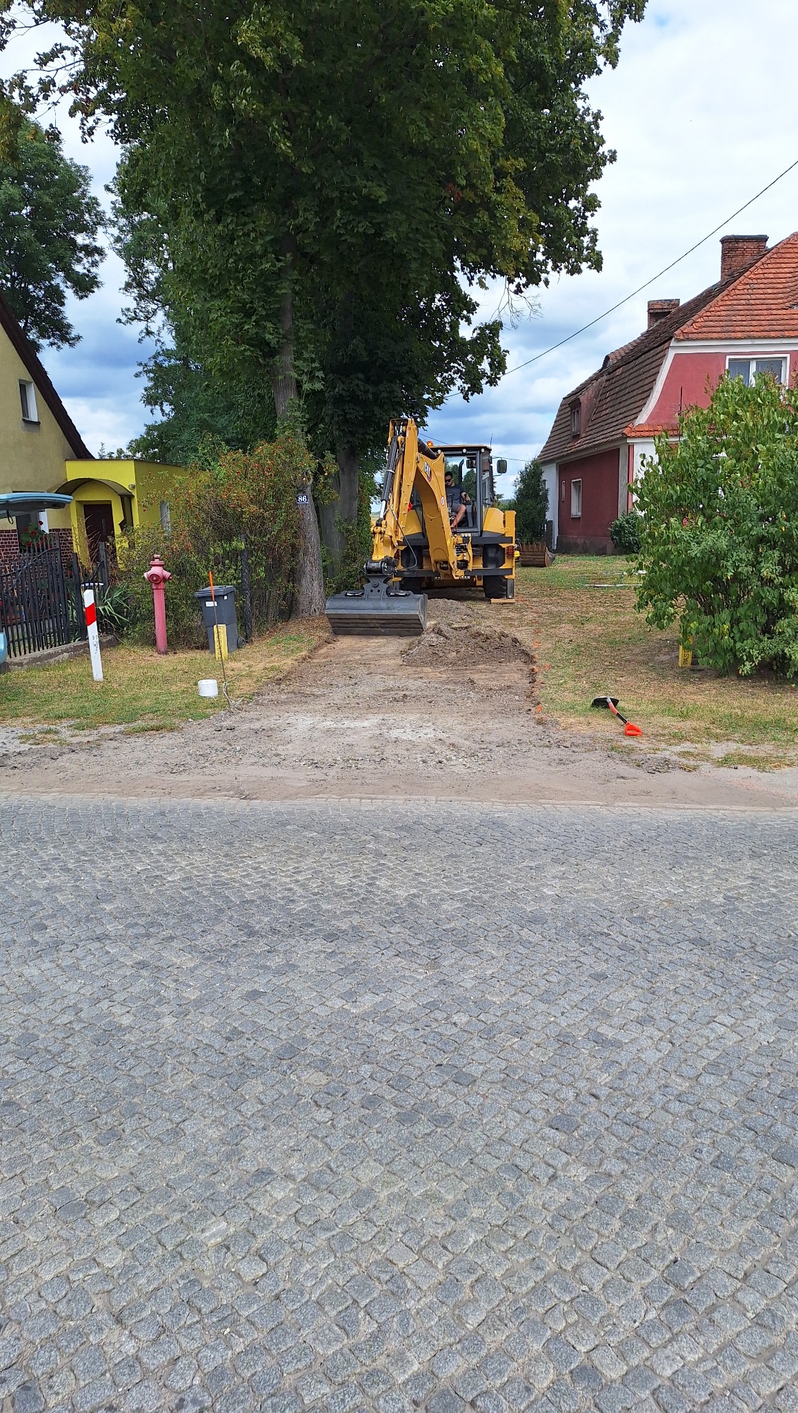 Żółta koparka na placu budowy, w tle domy i drzewa. Widoczny wykop pod drogę, pomarańczowa łopata i kostka brukowa. Etap prac ziemnych w wiejskiej scenerii.