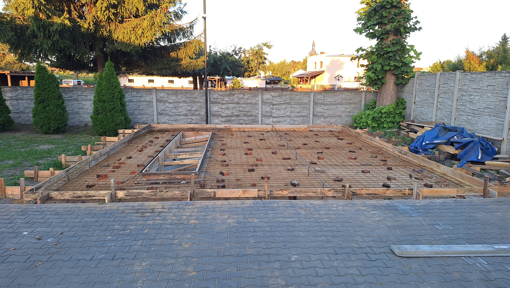 Zbrojenie fundamentów pod budynek, widoczne drewniane szalunki i siatka zbrojeniowa, cegły dystansowe. W tle ogrodzenie z betonu architektonicznego i zieleń.