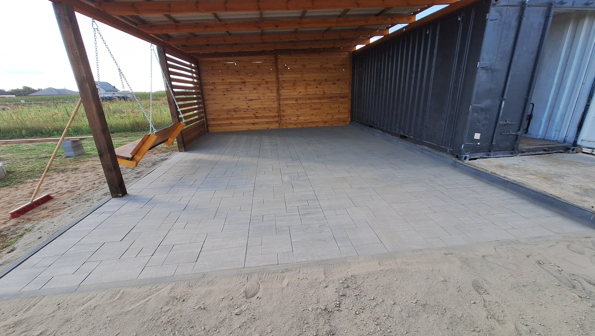 Ułożona szara kostka brukowa na posesji, przylegająca do metalowego kontenera i drewnianej wiaty z huśtawką. Widoczny piasek i częściowo trawa w tle.
