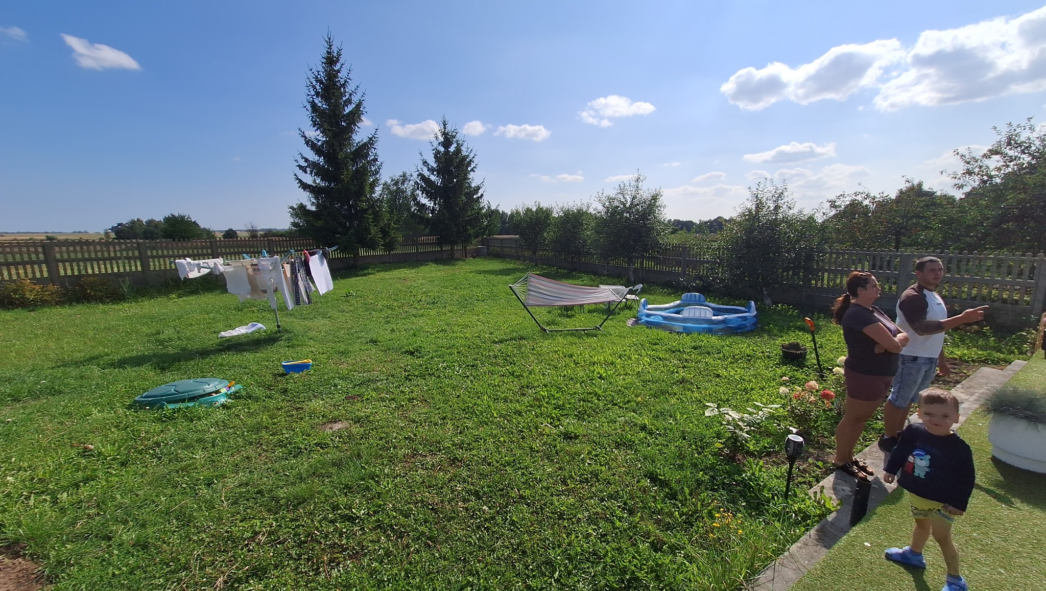 Słoneczny ogród z zieloną trawą, drzewami i domem w tle. Na pierwszym planie ludzie, zabawki, basenik i rozwieszone pranie. Widok na teren przed niwelacją.
