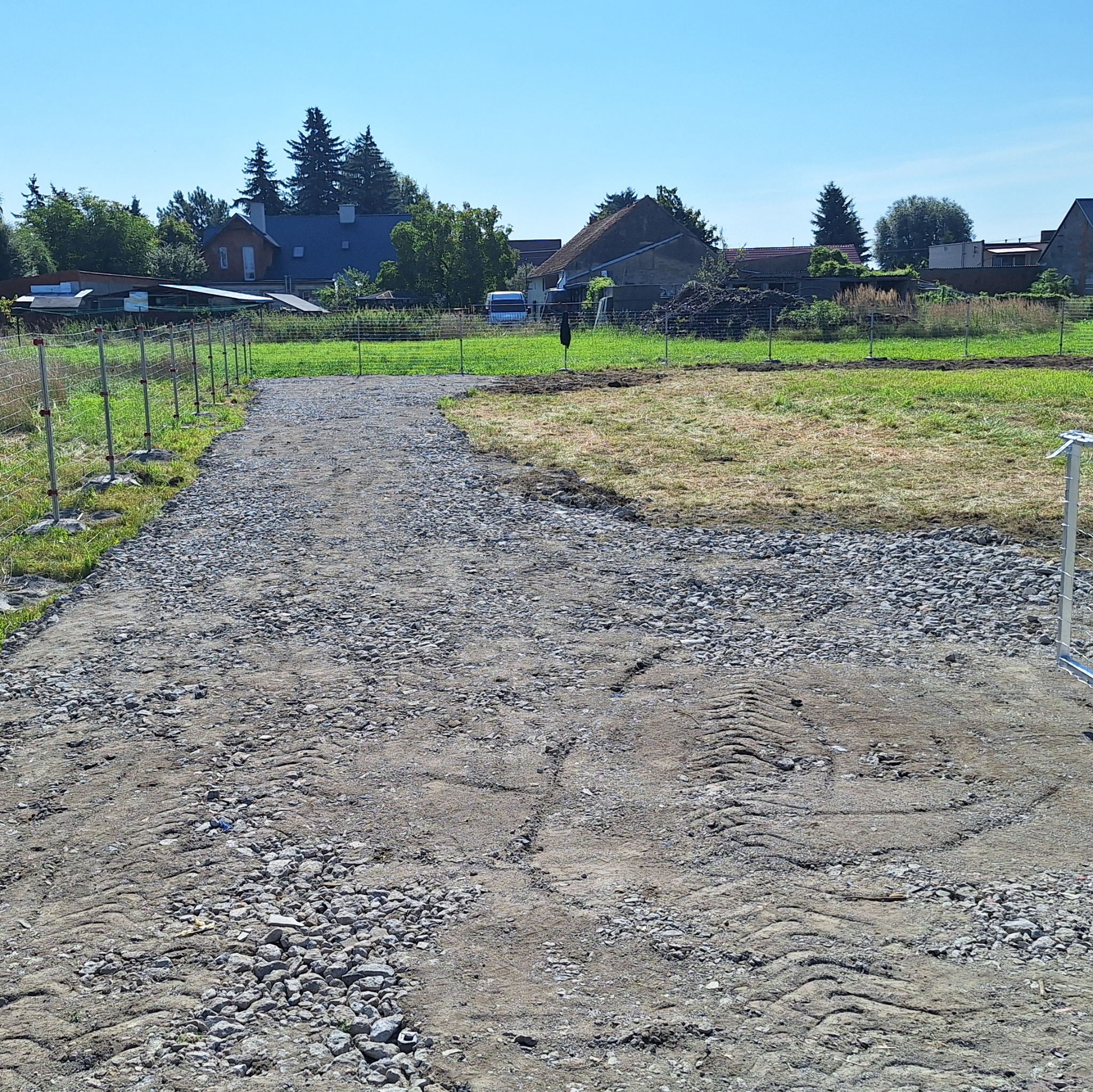 Utwardzona droga z szarego gruzu na działce, widoczne ślady opon. W tle domy i drzewa w letnim słońcu.