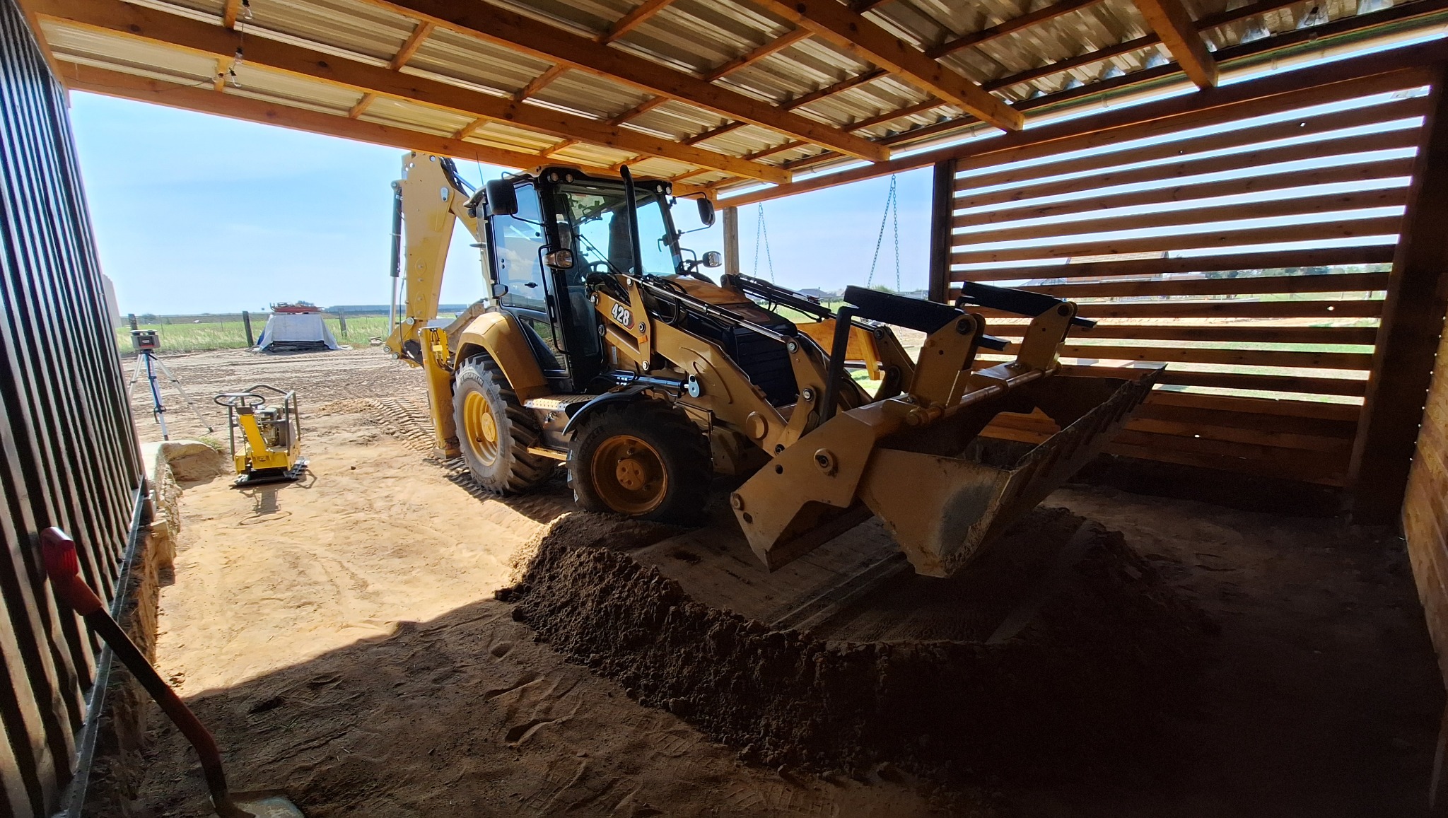 Żółta koparko-ładowarka CAT 428 w drewnianej wiacie, z wykopanym dołem i ubijarką w tle. Widoczny laser niwelacyjny, ziemia i drewniana konstrukcja.