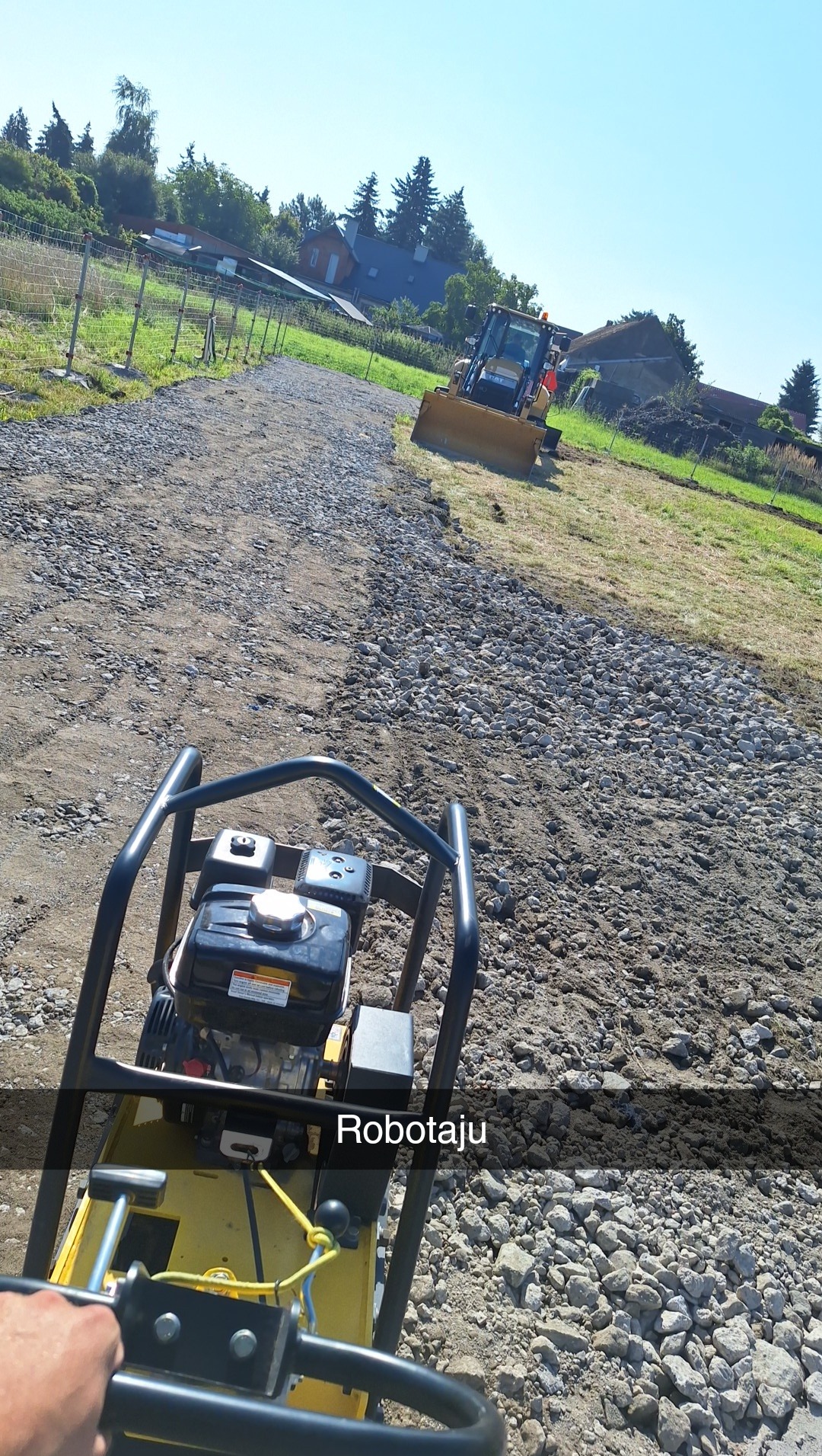 Praca zagęszczarką na żwirowej drodze; w tle spychacz na zielonym terenie, ogrodzenie i domy. Słoneczny dzień.