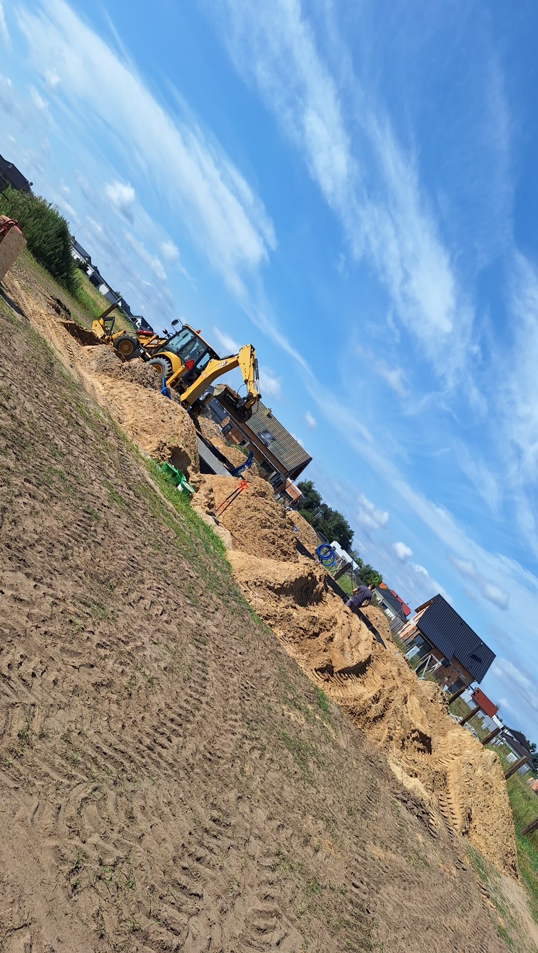 Żółta koparko-ładowarka przy wykopie fundamentów domu jednorodzinnego, widok z boku, w tle błękitne niebo z chmurami.