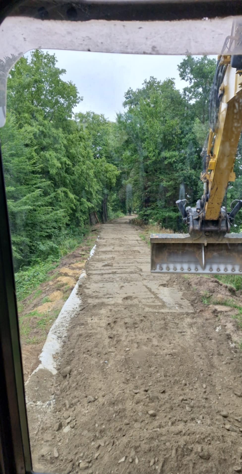 Widok z kabiny na utwardzaną drogę gruntową w lesie. Żółta łyżka koparki w kadrze. Obrzeża drogi zabezpieczone białym materiałem. Etap prac ziemnych.