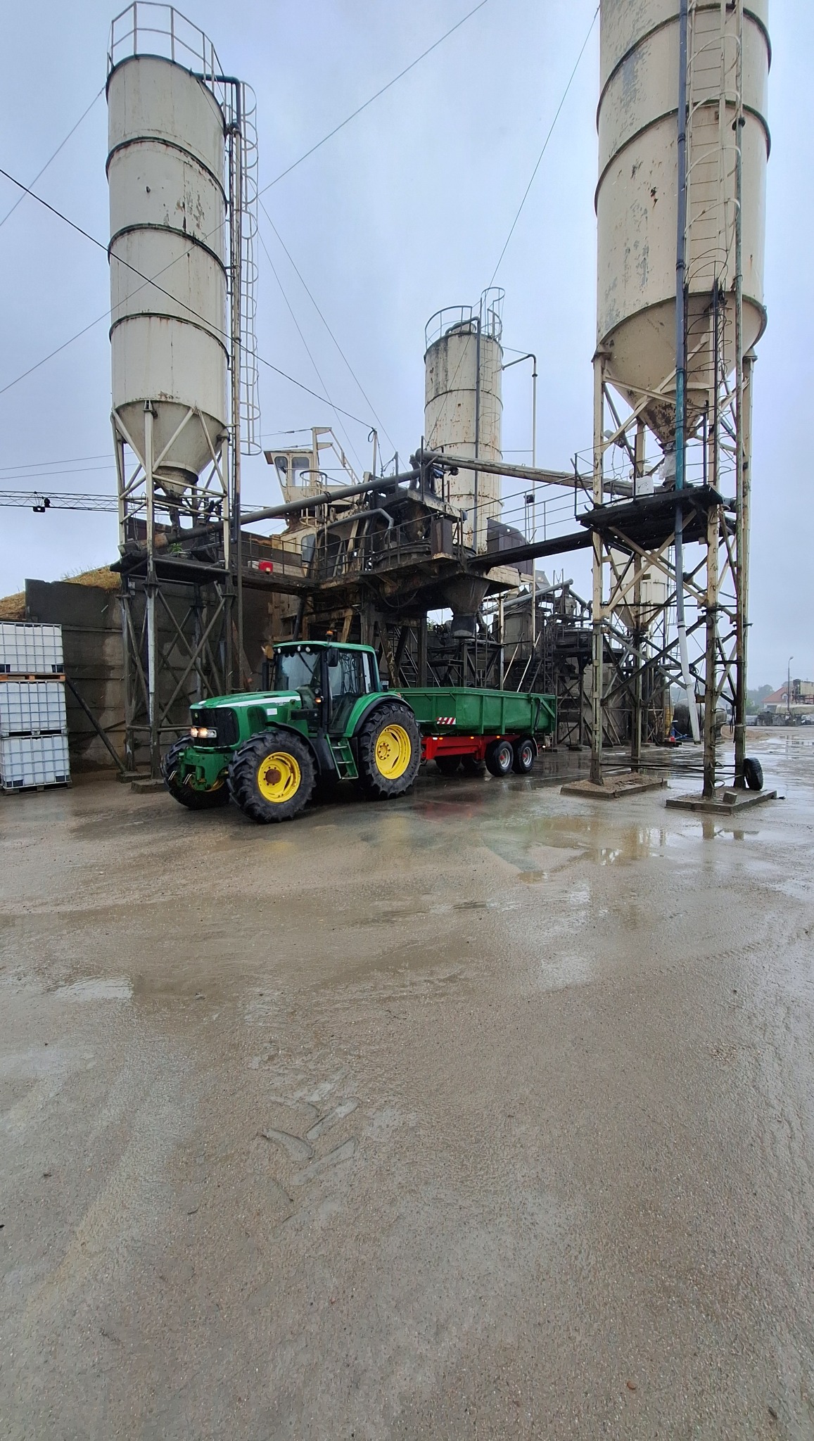Zielony traktor z przyczepą na mokrym terenie, w tle silosy i konstrukcja przemysłowa. Szary dzień, widoczne ślady opon na podłożu.
