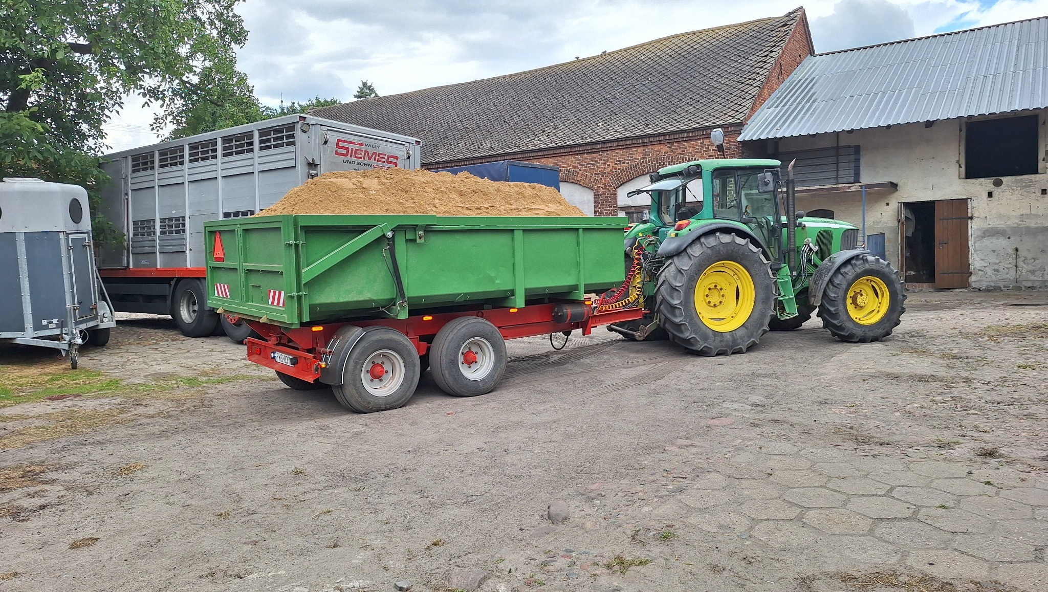 Zielony traktor John Deere z przyczepą wypełnioną piaskiem na placu, w tle budynki gospodarcze i przyczepy. Widoczne detale: mocowanie przyczepy, opony traktora.
