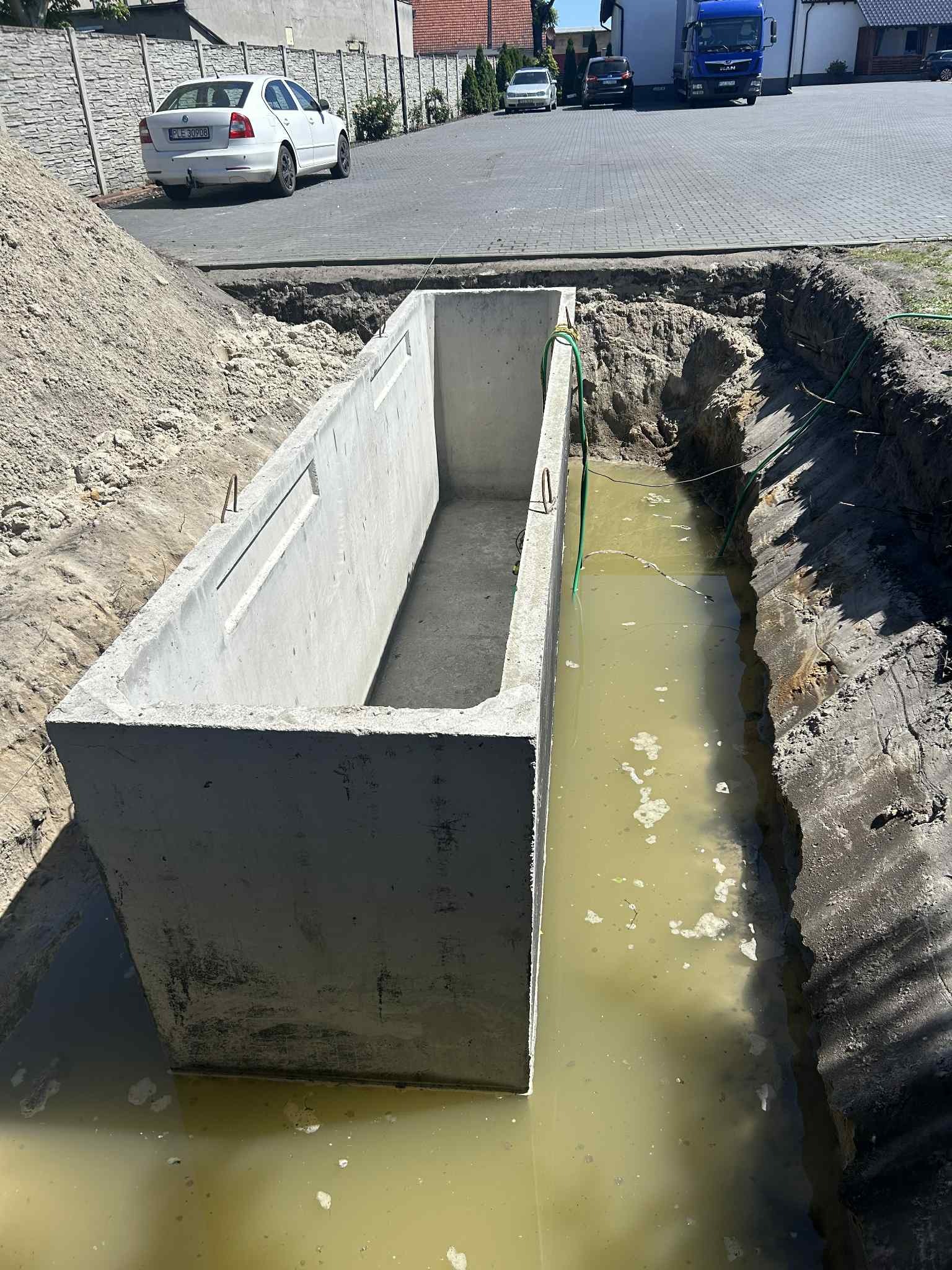 Betonowy zbiornik w wykopie wypełnionym wodą, widoczne ślady po wykopie, w tle samochody i budynki. Montaż szamba betonowego na działce.