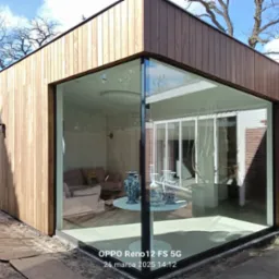 Nowoczesna, drewniana dobudówka z panoramicznym oknem narożnym, ukazująca jasne wnętrze z okrągłym stolikiem i dekoracyjnymi wazonami. Widoczne detale wykończenia.