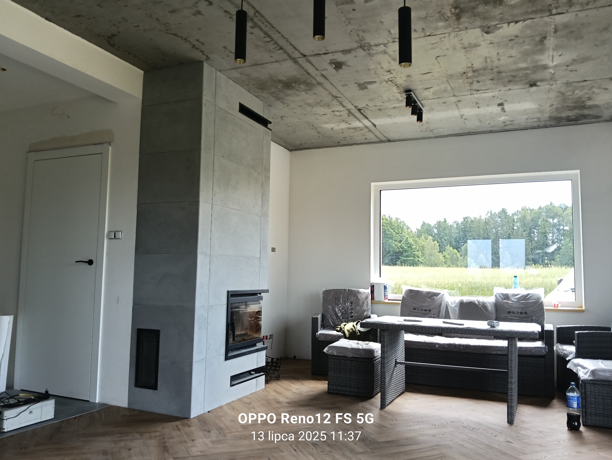 Nowoczesny salon z szarym kominkiem obłożonym płytami, drewnianą podłogą w jodełkę i widokiem na zieleń za oknem. Minimalistyczne lampy sufitowe.