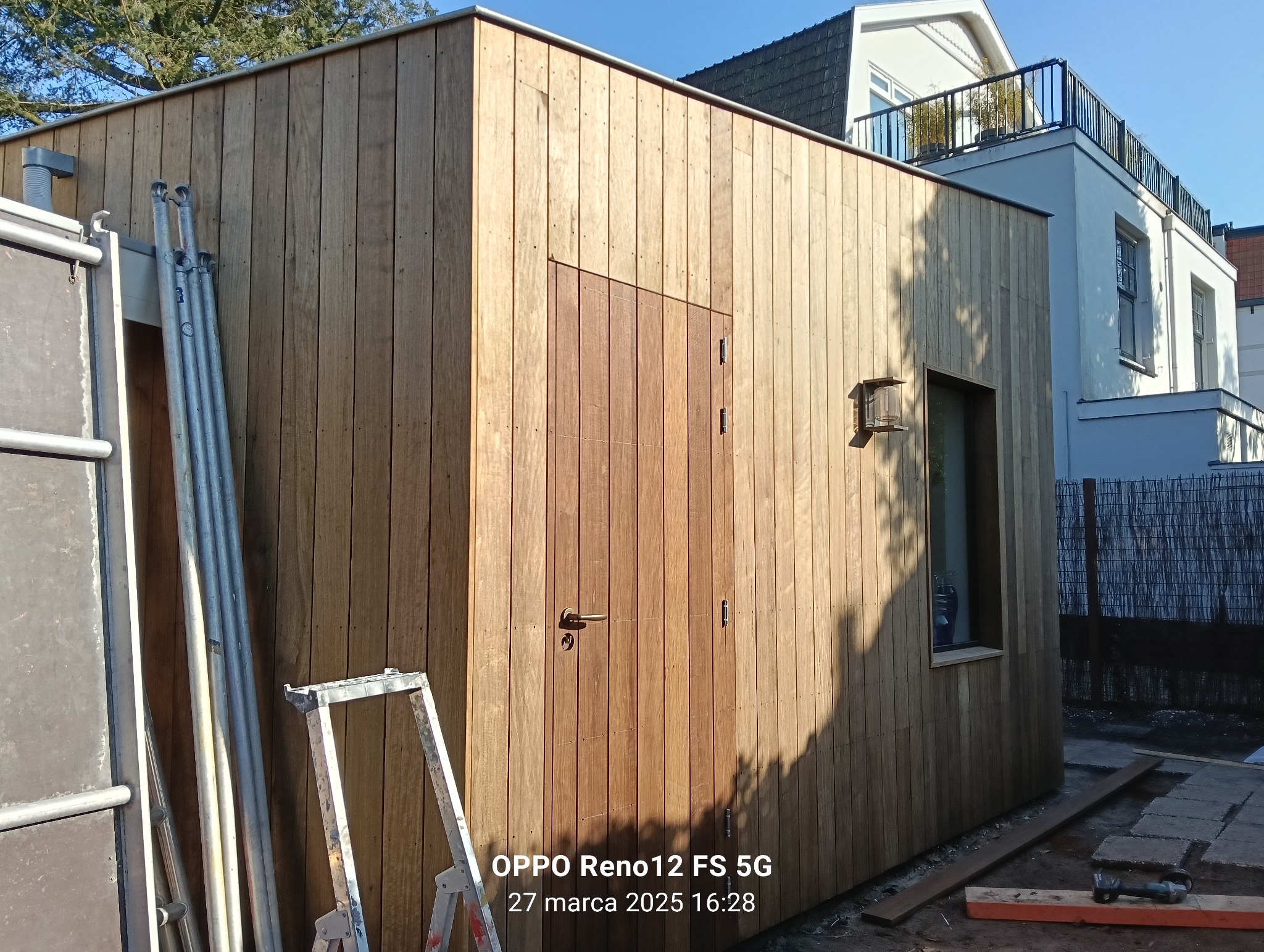 Nowoczesna, drewniana altana z oknem i drzwiami, na tle domu. Widoczne rusztowania i narzędzia sugerują prace wykończeniowe.