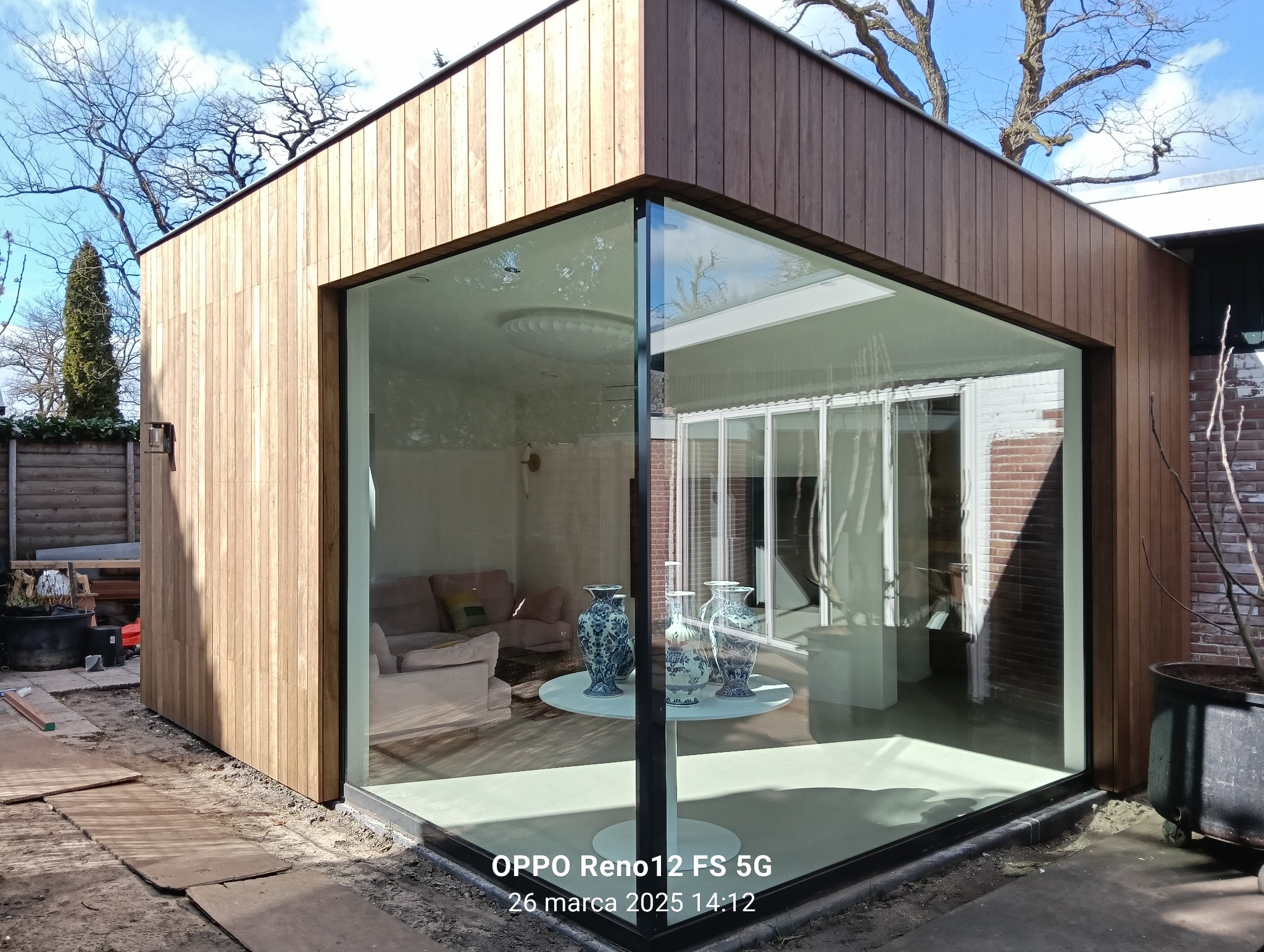 Nowoczesna, drewniana dobudówka z panoramicznym oknem narożnym, ukazująca jasne wnętrze z okrągłym stolikiem i dekoracyjnymi wazonami. Widoczne detale wykończenia.