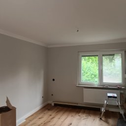 Remont4You - Pokój po malowaniu ścian na szaro z białą listwą sufitową i drewnianą podłogą. Widoczne drabina malarska i puszka z farbą. Okno z widokiem na zieleń.